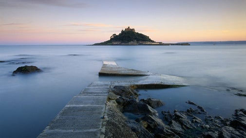 Silhouette view of St Michael's Mount from the causeway, with smooth sea and a pink dawn sky behind